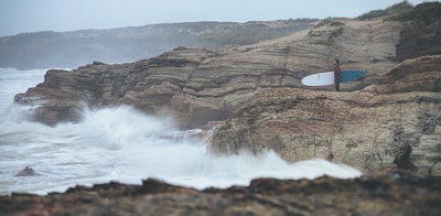 Standing on a mountain rocks during the day holding a white and blue surfboard to watch bodies of water
